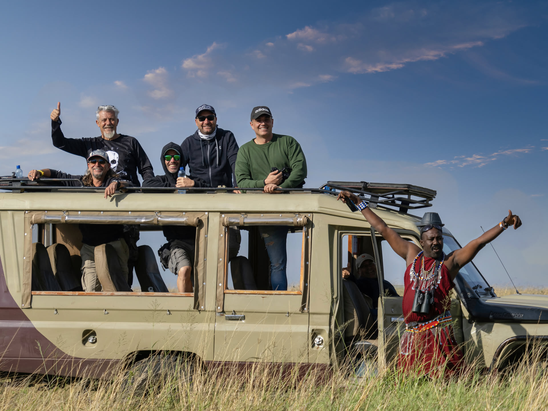 Grupo de viajeros de RAFIKI en un landcruiser de safari, saludando junto a un guía masái en vestimenta tradicional